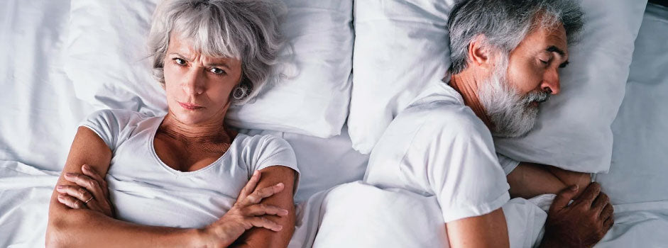 Femme senior éveillée et contrariée au lit près de son partenaire endormi — SANUSq Health